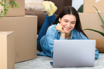 Moving to a new house, rental housing. Happy young caucasian woman using laptop computer to search and order a transportation service and movers to move to a new home - obrazy, fototapety, plakaty