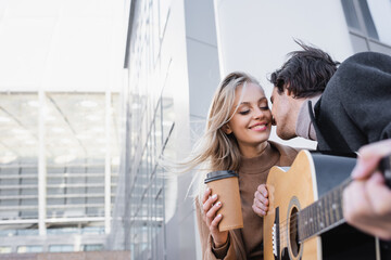 young man with acoustic guitar kissing happy blonde woman with coffee to go on city street.