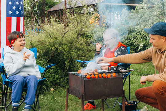 Happy young family make barbecue together in garden. People barbecuing meat on grill. Dad, son, daughter eating dinner lunch outdoors in backyard in summer on a sunny day. Leisure vacation holiday.