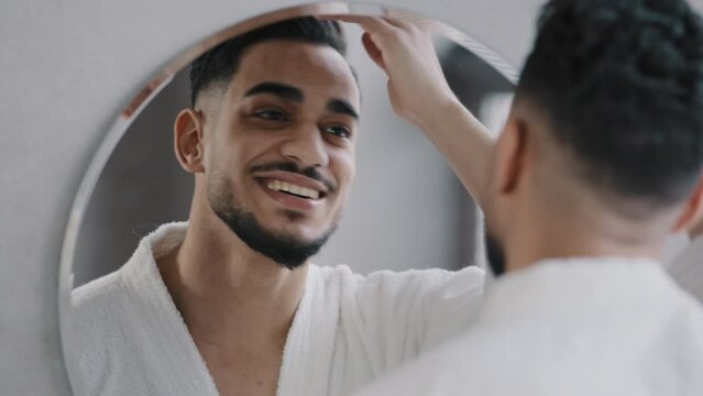 Handsome male model Indian hipster Arabian bearded guy doing hairstyle correct hair looking in mirror reflection. Smiling 30s man enjoying anti-dandruff shampoo result in morning grooming in bathroom.