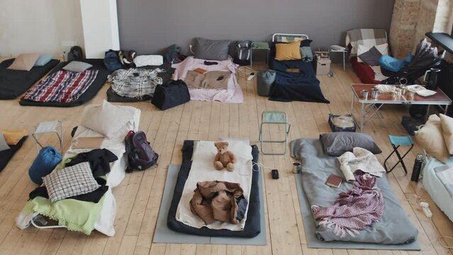 High angle of indoor refugee camp in afternoon, cots and mattresses with personal belongings and no people