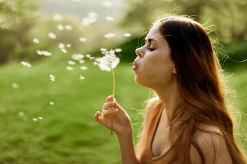 Woman with a dandelion flower in her hands smiling and blowing on it against a background of summer...