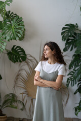 Portrait of calm brunette curly lady with natural make-up in summer outfit stand in cozy room with monstera background. Beautiful woman owner shop plants looking away, crossed hands. Gardening concept
