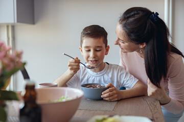 Portrait of small teen boy eating cereals with milk in kichen and mother looking with love.