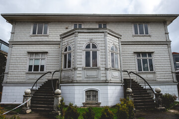 Front of a abandoned three story old centuries neoclassical white house with stone basement, two stairs and green garden in a cloudy day, Valdivia, Chile