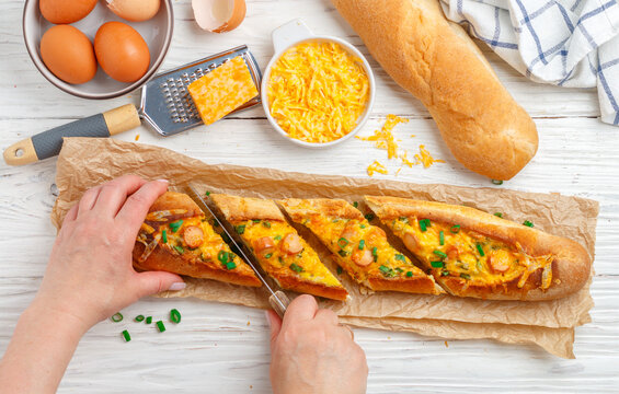Women's hands  cut  Baguette boat. Hot big sandwich in baguette bread with eggs, sausage, cheese and green onion on parchment on white wooden background. Breakfast. Selective focus. Top view