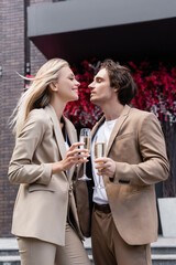 pleased couple in trendy suits holding champagne glasses and looking at each other on street.
