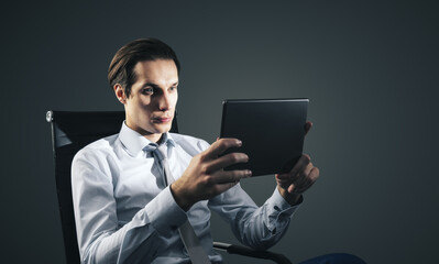 Young handsome businessman sitting on chair with digital tablet isolated on dark grey background