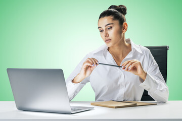 Online education and internet training concept with pretty young woman sitting at white table, looking at laptop screen and carrying pencil in hands on abstract light green wall background