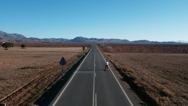 Inspiring aerial shot of millennial man ride on longboard in middle of empty road in american countryside. Summertime wanderlust inspiration. Freedom lifestyle of new generation