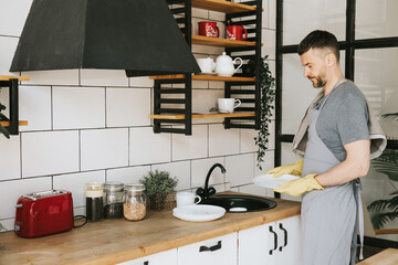 young man in apron and gloves does household chores, washes dishes with towel on his shoulder, men housework, household help in stylish kitchen in modern apartment