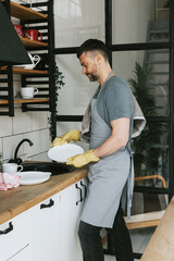 young man in apron and gloves does household chores, washes dishes with towel on his shoulder, men...