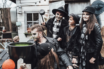 Obraz premium Scary family, mother, father, daughters celebrating halloween. Cauldron of potion.Terrifying black skull half-face makeup,witch costumes,stylish images.Horror,fun at children's party in barn on street