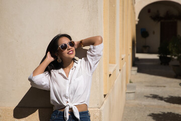 Portrait of beautiful young South American woman in white shirt, jeans and sunglasses on vacation in Europe lying on a wall sunbathing, touching her hair. Concept beauty, fashion, vacation, travel.