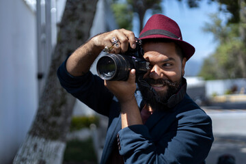 Obraz premium Latin American bohemian man with beard, hat and camera in hand taking pictures around the city. Career and vacation concept