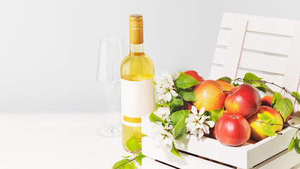 Bottle of apple wine with glass and farm apples in a white wooden box on a light neutral background with copy space. Summer Low-alcohol fruit drink. Fruit wine. Apple product. Soft focus style