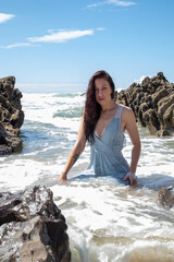 A woman in a blue dress at the beach on a sunny day. The blue sky has some cloud cover. The model sits amongst the rocks as the sea washes waves over her