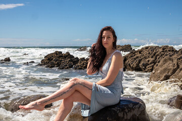 Fototapeta na wymiar A woman in a blue dress at the beach on a sunny day. The blue sky has some cloud cover. The model sits amongst the rocks as the sea washes waves over her