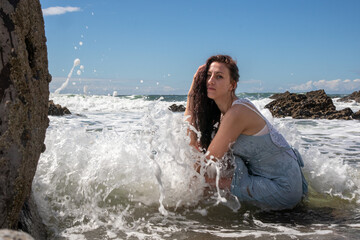 A woman in a blue dress at the beach on a sunny day. The blue sky has some cloud cover. The model sits amongst the rocks as the sea washes waves over her