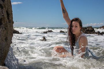 A woman in a blue dress at the beach on a sunny day. The blue sky has some cloud cover. The model sits amongst the rocks as the sea washes waves over her