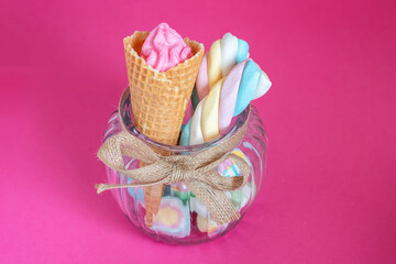 Candies and marshmallows with waffle cone in glass jar on pink background. Sweets, sugar, candies, marshmallow and ice cream cone. Free copyspace, pastel colours.