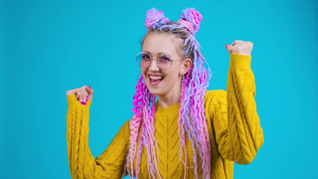 Funny young woman with original style of hair dancing in studio on blue background. She has colorful clothes, afro braids dreadlocks. Happy person moving, positive emotions, ecstatic feeling