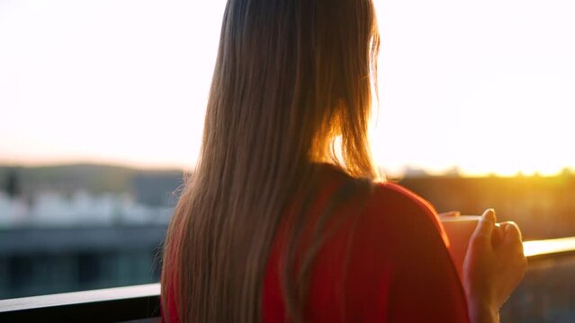 Woman drinking coffee while standing on the balcony and admire the sunset. Slow motion