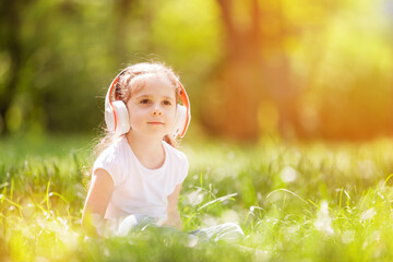 Cute little girl is listening to music in the spring park. Family summer outdoor lifestyle. Happy small kid in headphones sitting on green grass. Beauty nature at summer. Childhood happiness - 506889994