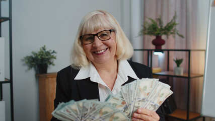 Senior mature banker business woman accountant wears glasses, formal suit, celebrating business success dancing with stack of money dollar cash. Elderly grandmother freelancer indoors office interior