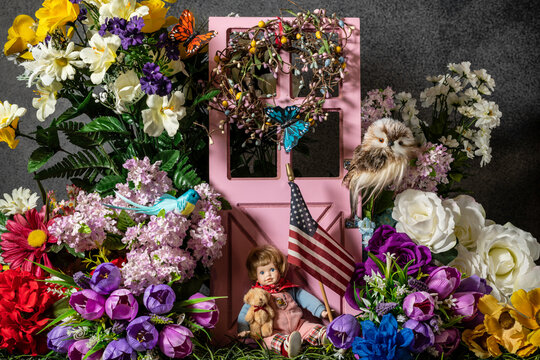 child doll sitting on grass holding American flag and teddy bear surrounded by flowers