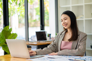 Asian businesswoman analyzing a report pointing to a graph with a pen, laptop, and calculator In her workstation, accountant, telemarketing ideas, e-commerce, free online marketing.