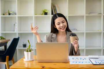 Smiling beautiful Asian businesswoman holding coffee cup and laptop working on graph papers at financial accounting concept office