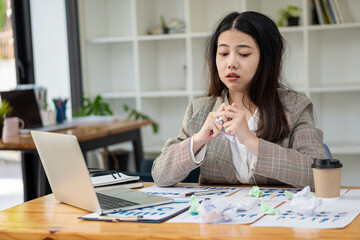 Asian businesswoman with work stress crumples paper, financial graph document on laptop table at financial accounting concept office.