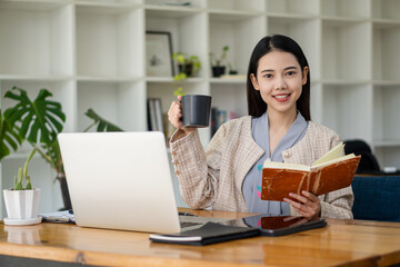 A beautiful Asian businesswoman is drinking coffee and working on a laptop with a tiger skin notebook on her desk at the office.