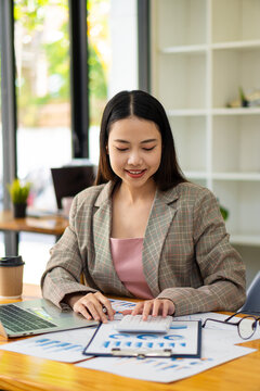 Smiling beautiful Asian businesswoman sitting with laptop and computer working on a financial graph document at the office. financial accounting concept vertical picture