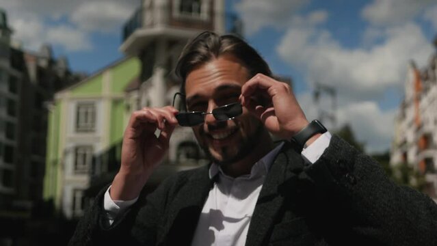 Cheerful smiling guy putting sunglasses on outside and shaking head affirmatively. Slow-motion of beaming handsome male wearing shades on a bright sunny day. Attractive happy guy gladly grinning
