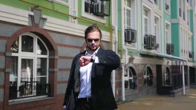 Confident handsome caucasian man walking on street and checking watch in slow motion. Successful rich male looking at watch on his way to earn more money. Wealthy guy in sunglasses checks wristwatch 
