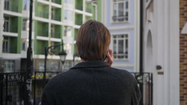 Camera following caucasian man talking on phone in slow motion. Back view of brown-haired male walking and discussing news on cell phone. Footage behind guy with stylish haircut conversing on phone
