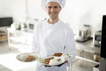 Portrait of young handsome smiling chef in uniform standing with ready meals in the kitchen. Happy caucasian cook working at restaurant. Concept of professional occupation and haute cuisine
