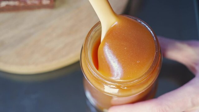 close-up white spoon stirs a full jar of liquid homemade salted caramel. viscous, tasty, appetizing caramel flows down the spoon. homemade sweets
