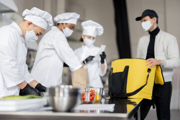 Courier waiting for an order for delivery in the kitchen with chefs preparing takeaway food. Concept of dark kitchen and delivering food during pandemic. Multiracial cook team in face mask and