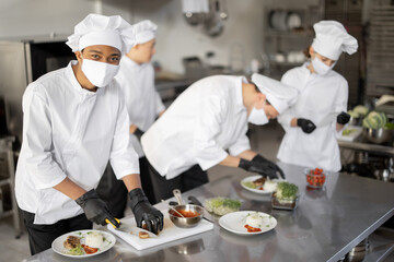 Multiracial group of cooks finishing main courses while working together in the kitchen. Cooks wearing uniform and face mask. Team prepares meals for the restaurant