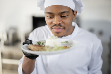 Portrait of young handsome smiling chef in uniform standing with ready meal in the kitchen. Happy Latin-American cook working at restaurant. Concept of professional occupation and haute cuisine