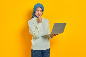 Pensive beautiful Asian woman in white sweater using laptop looks seriously thinking about something isolated over yellow background. people religious lifestyle concept