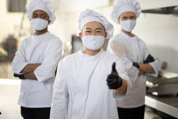 Portrait of three well-dressed chef cooks with different ethnicities standing together in restaurant kitchen. Asian chef showing OK sign, latin and european guys on background. Cooks wearing face