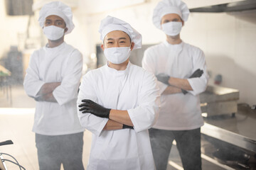 Portrait of three well-dressed chef cooks with different ethnicities standing together in restaurant kitchen. Asian chef standing in front, latin and european guys on background. Cooks wearing face