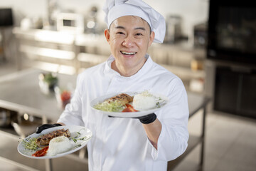 Portrait of young happy handsome smiling chef in uniform standing with ready meal in the kitchen. Happy asian cook working at restaurant. Concept of professional occupation and haute cuisine
