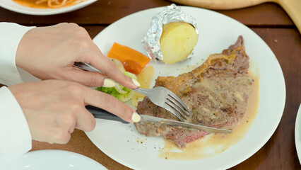 two beautiful hands holding a knife and fork, slicing a large piece of steak in an appetizing dish, mashed potatoes and vegetables on a plate.