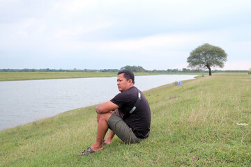 An Indonesian young man sitting by the lake with a sling bag over his shoulder