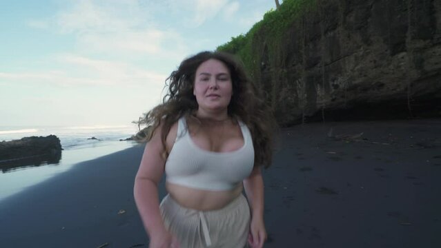 Happy plus size girl with long hair running on the black sand beach. Body positivity. 4K
